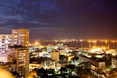 Kinshasa by night showcases a city illuminated by countless lights, with a bustling port and towering buildings reflecting in the calm waters of the Congo River under a twilight sky.